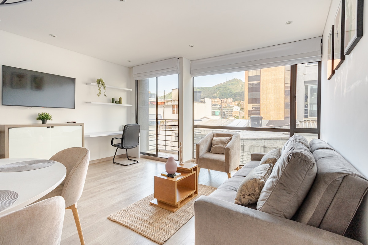 A modern living area features large windows providing natural light and mountain views. A comfortable sofa is positioned beside a wooden coffee table, with a decorative item placed on it. A dining area with a round table and upholstered chairs complements the stylish décor.