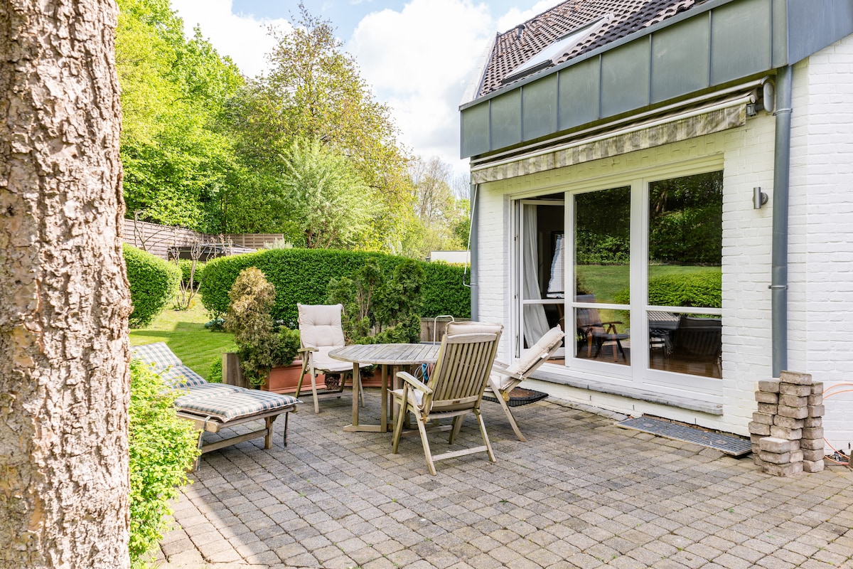 An outdoor patio features a mix of seating options, including a dining table with chairs and a lounge chair. Lush greenery surrounds the space, providing a serene atmosphere. The patio is paved with stone, offering ample room for relaxation and enjoyment.