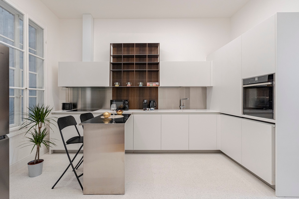 A modern kitchen is shown, featuring sleek white cabinetry and a contrasting wooden shelving unit. An island counter with two black stools creates a functional dining space, while high-end appliances are integrated into the design. A small potted plant adds a touch of greenery.
