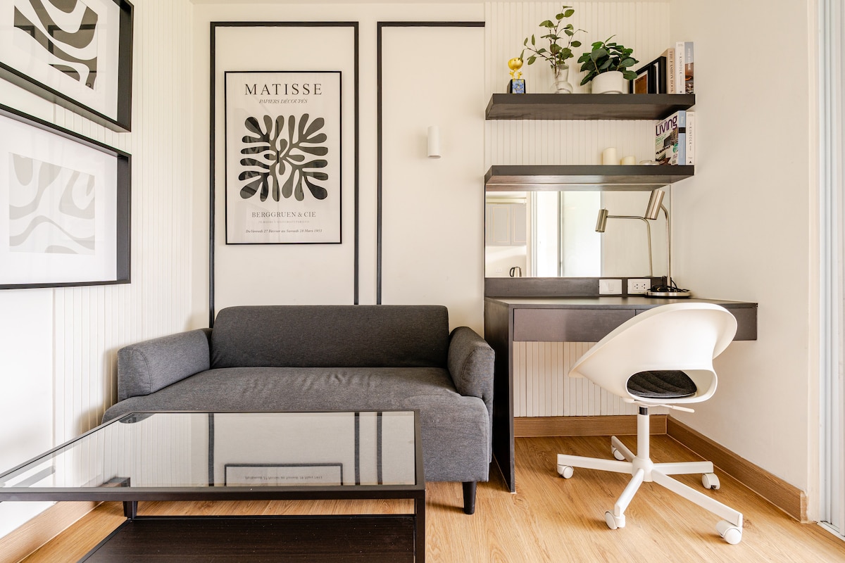 A compact living area is presented, featuring a gray sofa adjacent to a glass coffee table. A wall-mounted workspace, including a desk and chair, is positioned beside a decorative plant, with wall art providing a modern touch. Natural light enters through a nearby window.