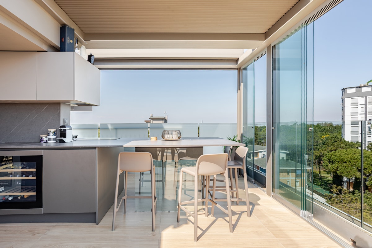 A modern kitchen area features sleek cabinetry and high-quality appliances. A spacious dining table with light-colored chairs is positioned near glass walls, allowing for unobstructed views of the sea and surrounding nature. Natural light floods the space, enhancing the airy feel.