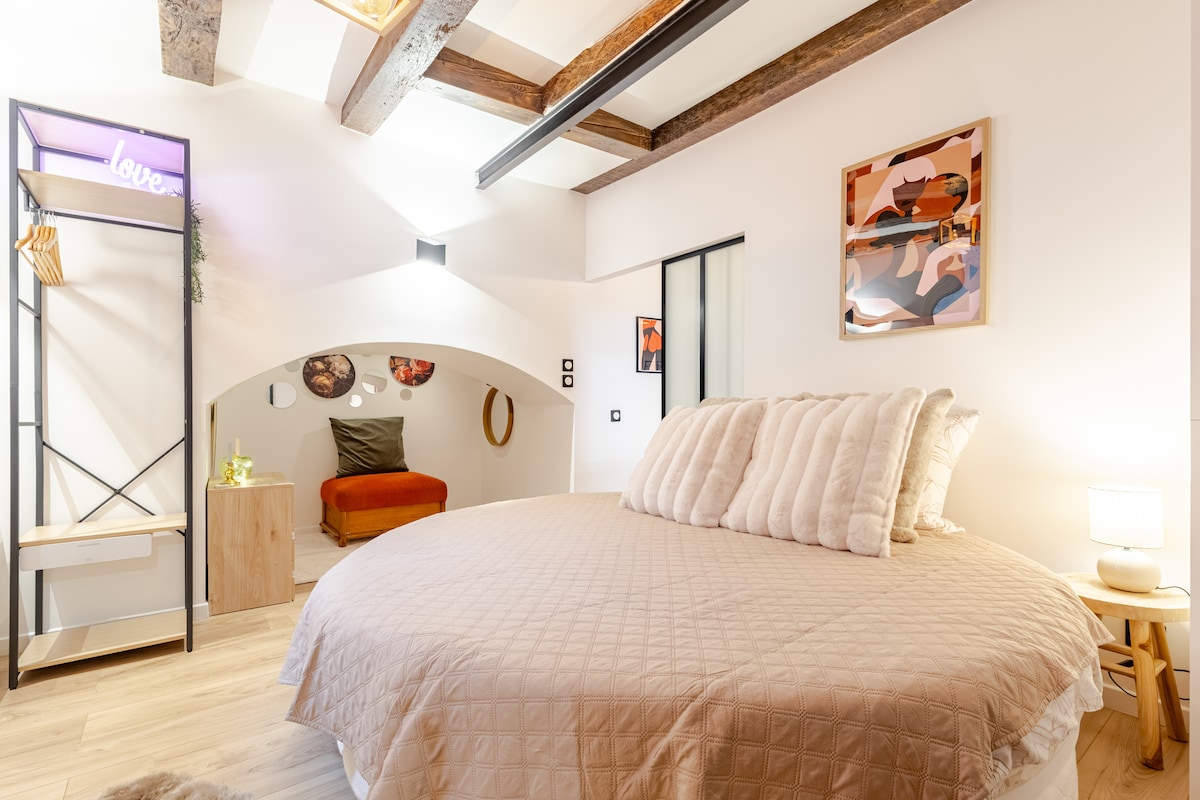 A spacious room features a large round bed with a textured quilt, complemented by wall art and decorative elements. Exposed wooden beams add character to the ceiling. A cozy seating area is visible to the side, enhancing the intimate atmosphere.