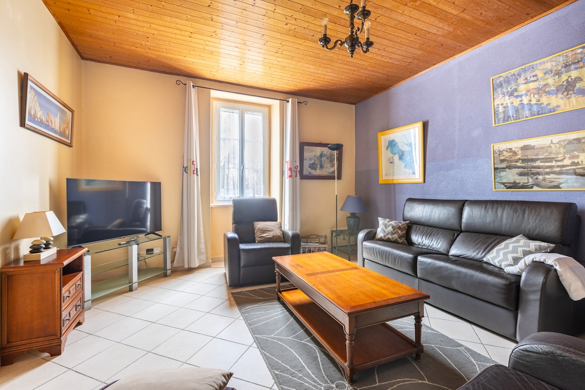 A comfortable living area features a dark leather couch paired with a wooden coffee table. A flat-screen television is positioned near a window, allowing natural light to enter the room. The walls are adorned with framed artwork, and warm wooden accents enhance the ambiance.