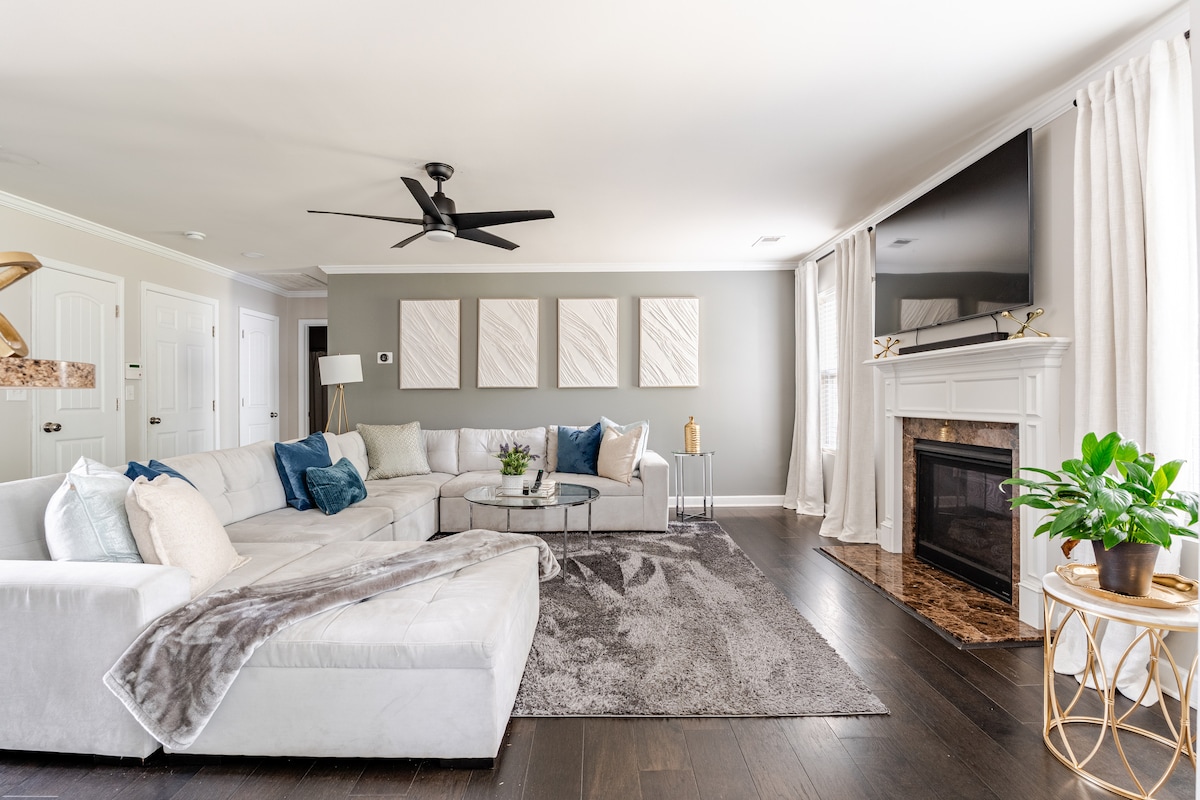 A spacious living room is featured with a sectional sofa adorned with pillows, accompanied by an area rug and a coffee table. Large windows draped with light curtains allow natural light to fill the room, where a fireplace is set against a neutral wall, complemented by decorative wall art.