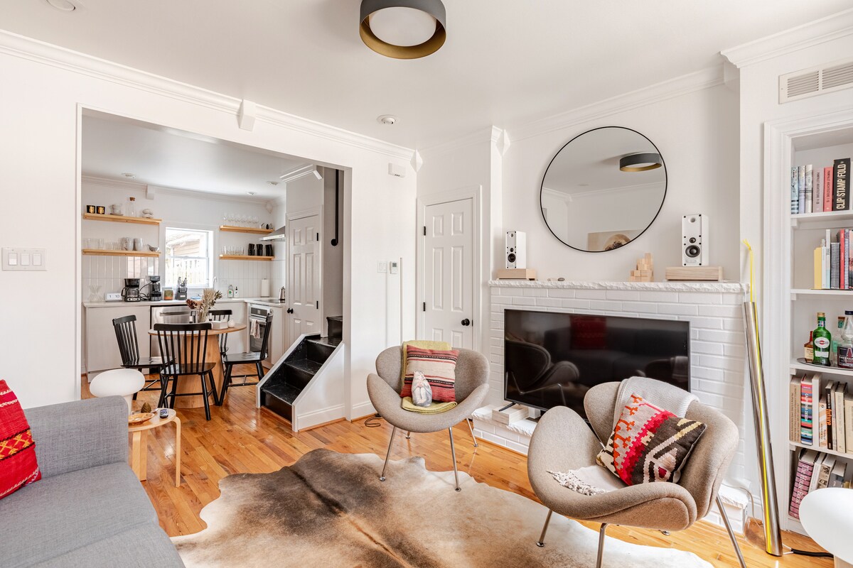 A cozy living area features a gray couch and two contemporary armchairs, arranged around a low coffee table on a plush cowhide rug. A sleek wall-mounted TV sits above a white brick fireplace. A mirror enhances the natural light from the nearby kitchen area.