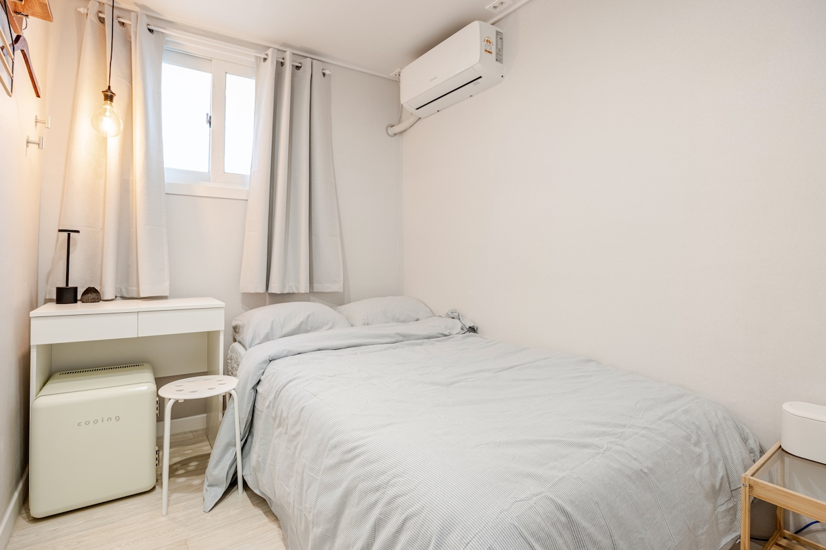 A clean and minimalistic single room is displayed, featuring a spacious bed with soft bedding. A small desk is positioned beside the bed, accompanied by a compact stool. A window with light curtains allows natural light to enter the room, while an air conditioning unit is mounted on the wall.