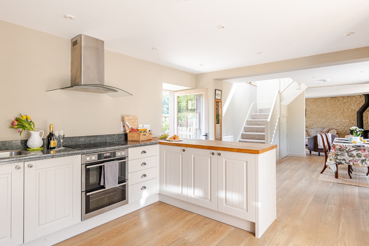 The spacious kitchen features a light-coloured cabinetry with a wooden countertop, complemented by modern appliances including a double electric oven and induction hob. A window allows natural light to flood the area, enhancing the inviting atmosphere. Fresh flowers add a touch of colour to the decor.