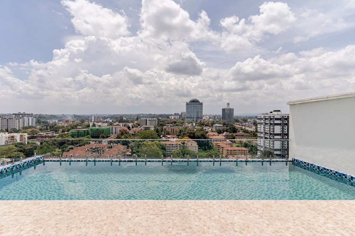 A rooftop swimming pool offers serene views of the city skyline, with lush greenery and modern buildings in the distance. The pool's clear water reflects the clouds above, creating a tranquil atmosphere against the backdrop of urban life.