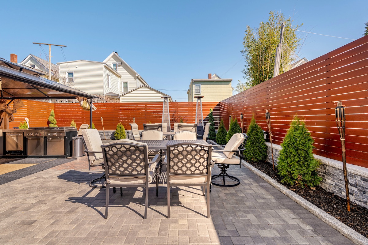 A spacious outdoor patio area is presented, featuring several seating options arranged around a central table. Lush greenery lines the perimeter, while a modern barbecue setup is visible in one corner. A wooden fence ensures privacy and creates a welcoming space.