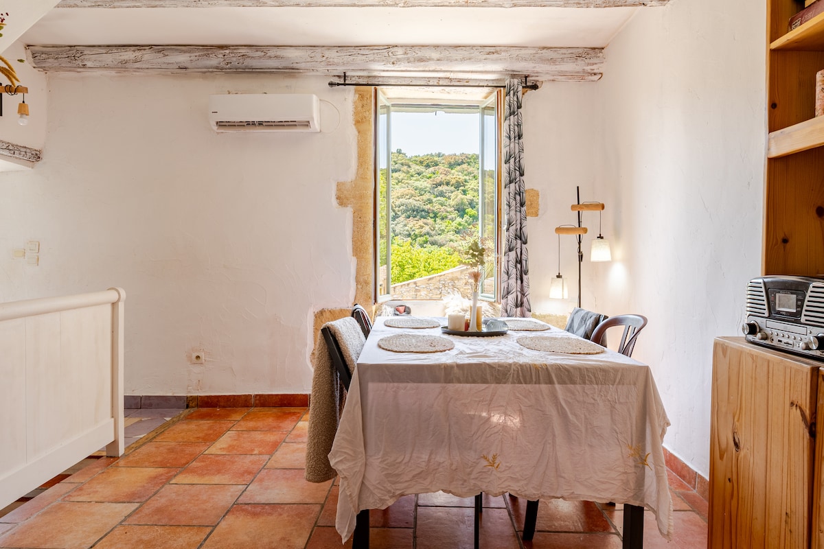 A dining area is presented with a wooden table set for four, featuring a light tablecloth. Natural light enters through a window, showcasing views of the surrounding greenery. A vintage radio and wooden shelving add to the rustic charm of the space.