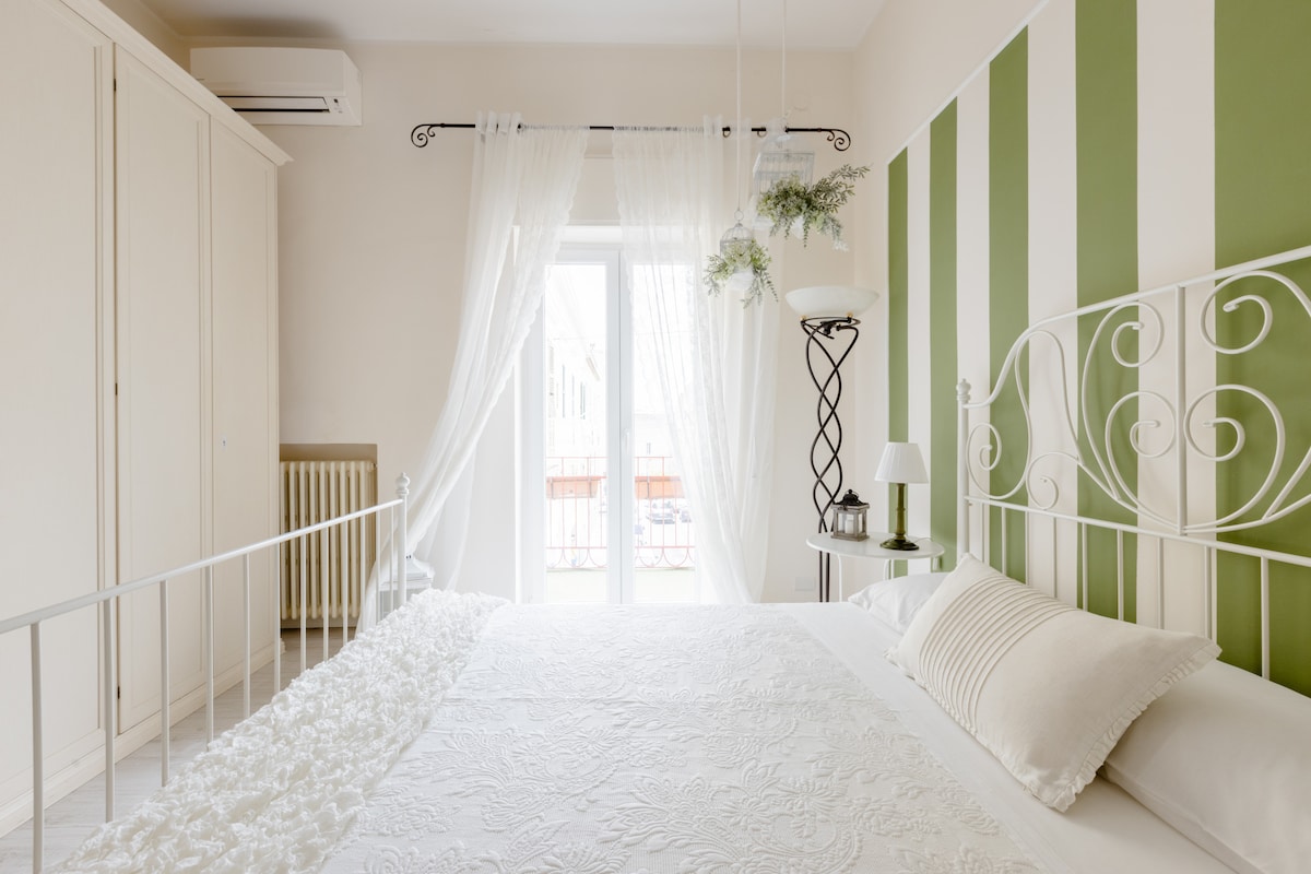 A bright and inviting bedroom features a white iron bed with a textured bedspread. Striped green and white walls complement soft lighting from the bedside lamp. Sheer curtains frame the balcony door, allowing natural light to fill the space. A wardrobe and radiator are neatly positioned against the wall.