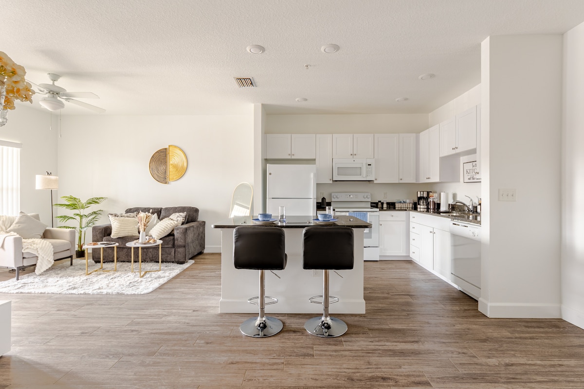 A modern kitchen and living area are connected in an open layout. The kitchen showcases white cabinetry, a stove, and a dishwasher next to a compact dining island with two black bar stools. A cozy seating arrangement on the side includes a grey sofa and green plants.