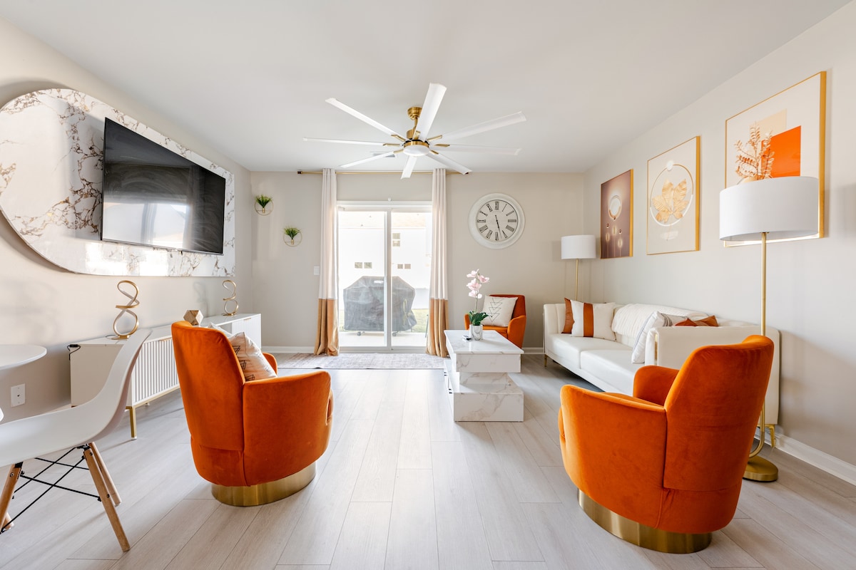 A spacious living area features a modern design with two vibrant orange armchairs and a sleek white sofa. A large wall-mounted TV is displayed above a marble accent piece. Natural light enters through glass doors, leading to an outdoor area, complemented by elegant wall art.