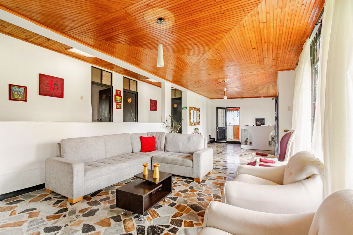A spacious living area is characterized by wooden beams overhead and natural stone flooring. A large gray sectional sofa complements a dark coffee table, placed centrally. Light-colored chairs are positioned nearby, with large windows allowing ample natural light to fill the space.