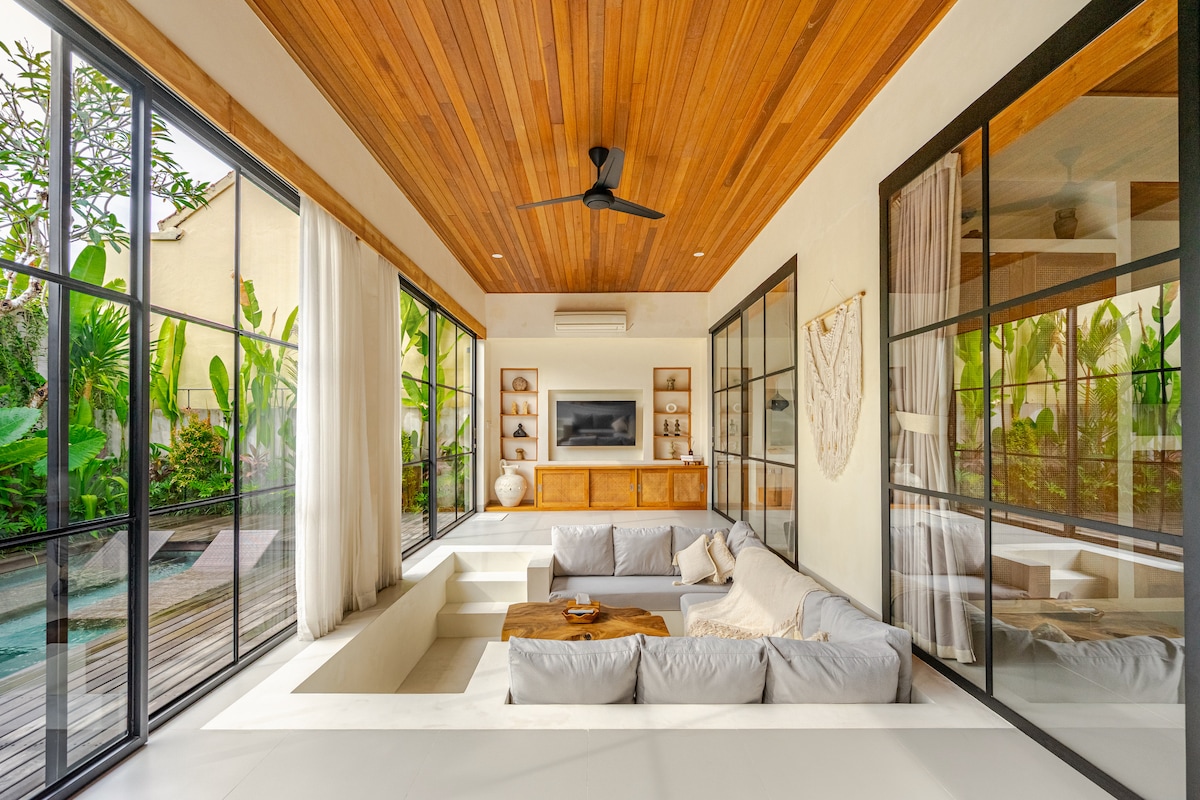 A spacious living area features large glass doors that allow natural light to fill the room. A sunken sofa is arranged around a wooden table, and a mounted TV is visible on the wall. Tropical plants are seen through the glass, enhancing the serene environment.