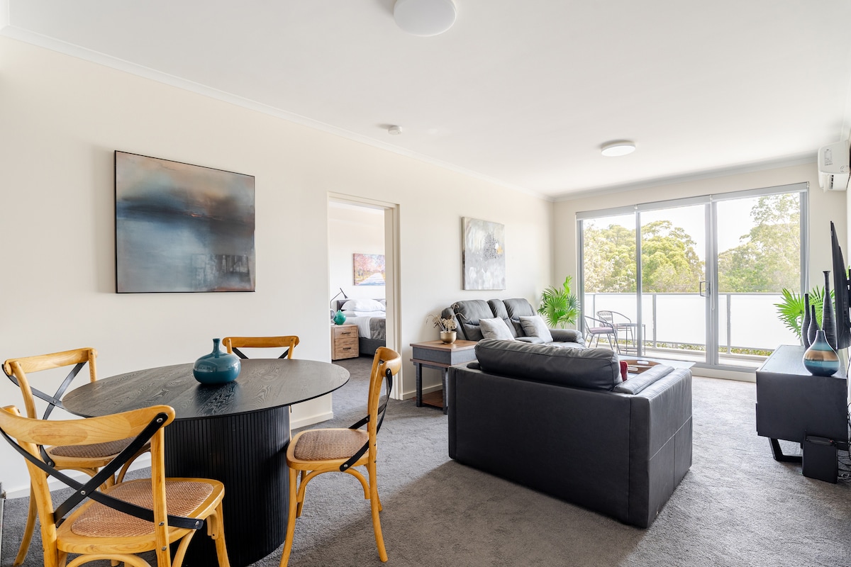 A living area is shown with a dark leather sofa, a round dining table with four chairs, and wall art. Natural light filters through large glass doors that open to an outdoor space, with leafy greenery visible in the background.