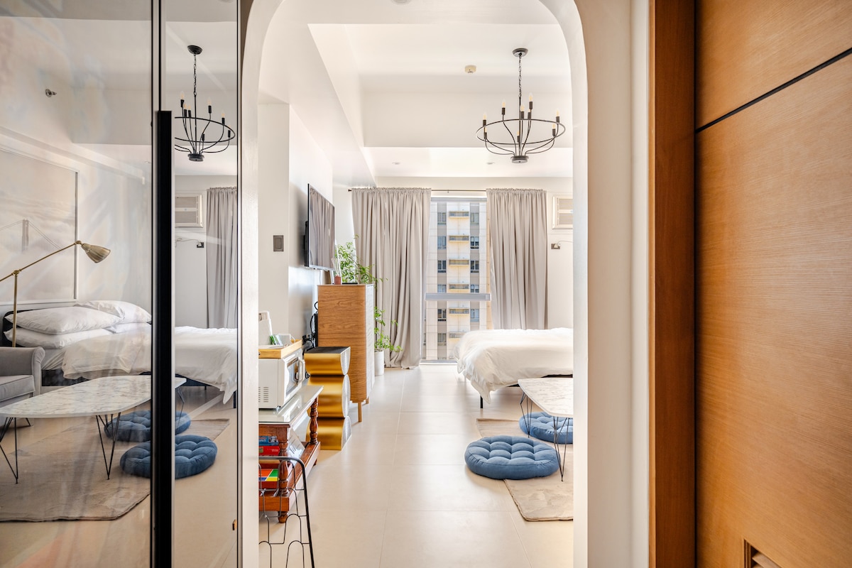 A spacious studio unit is revealed through an arched doorway. Light-colored walls and soft curtains create an airy atmosphere. A queen-sized bed is visible alongside a lounging area with a low table and floor cushions. A glimpse of a modern entertainment setup adds to the inviting feel.