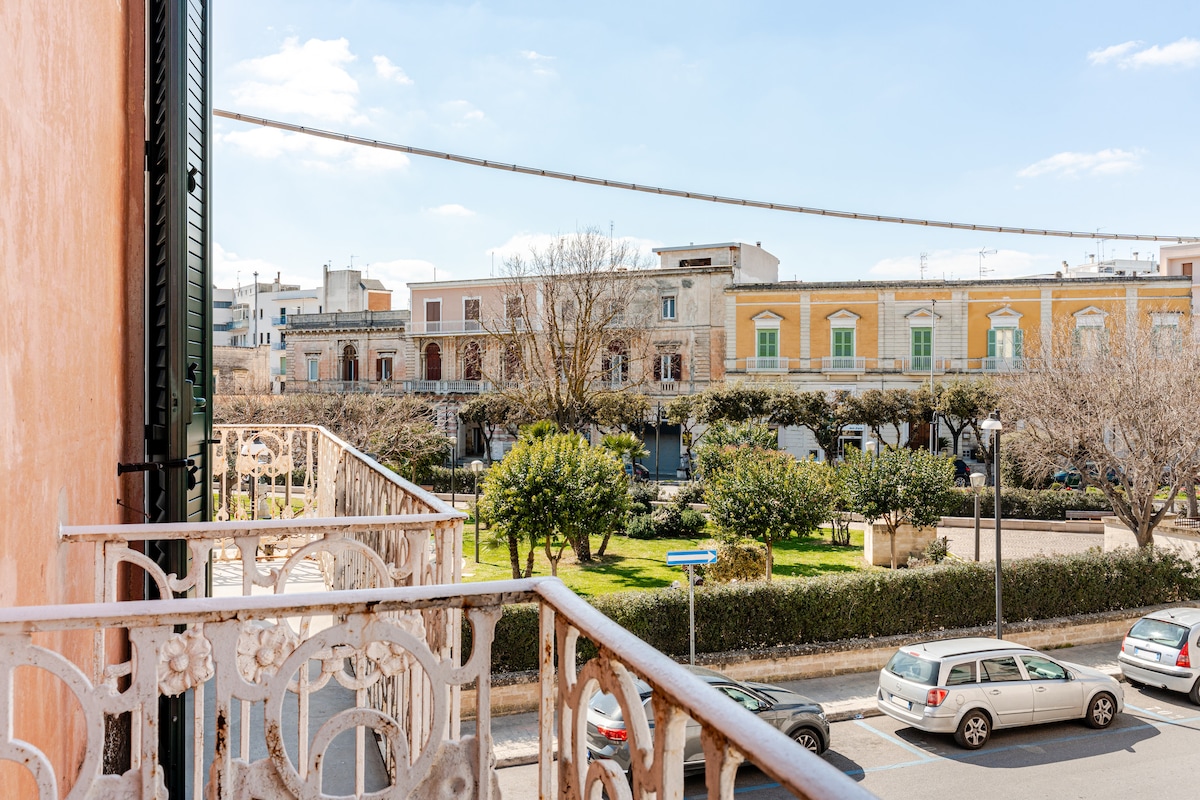 A view from a balcony showcases a well-maintained park and tree-lined streets. Colorful building facades can be seen in the background, while parked cars line the road below. The scene is illuminated by natural light, providing a sense of openness and calm.