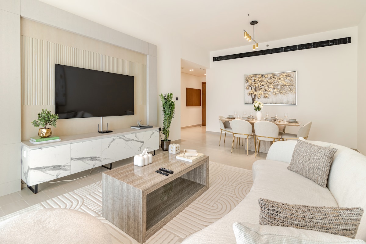 A modern living space is presented, featuring a large flat-screen television mounted above a sleek marble-accented console. A comfortable sofa invites relaxation, while a dining area with elegant chairs and a minimalist table is visible in the background, complemented by soft lighting.
