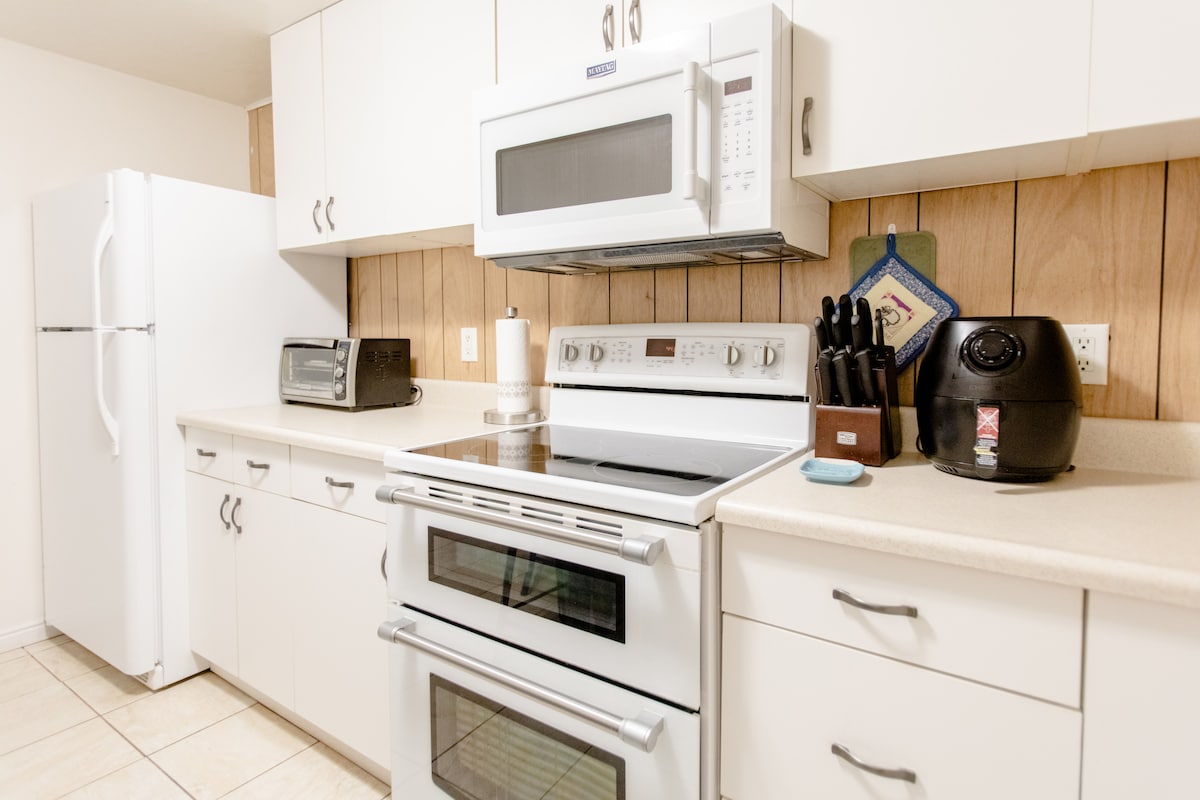 A well-equipped kitchen features modern white cabinetry, highlighted by a microwave and stove. An adjacent refrigerator and a toaster oven are positioned on the countertop. A set of knives is arranged neatly in a black holder, with a rice cooker visible beside it.