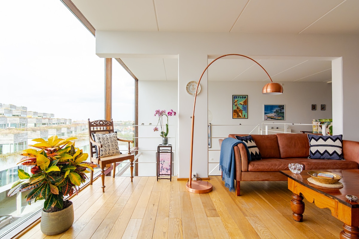 A spacious living area features a brown sofa, an elegant floor lamp, and a unique coffee table. A vibrant plant sits nearby, while artwork adorns the walls. Floor-to-ceiling windows allow natural light to fill the space, revealing views of the surroundings.