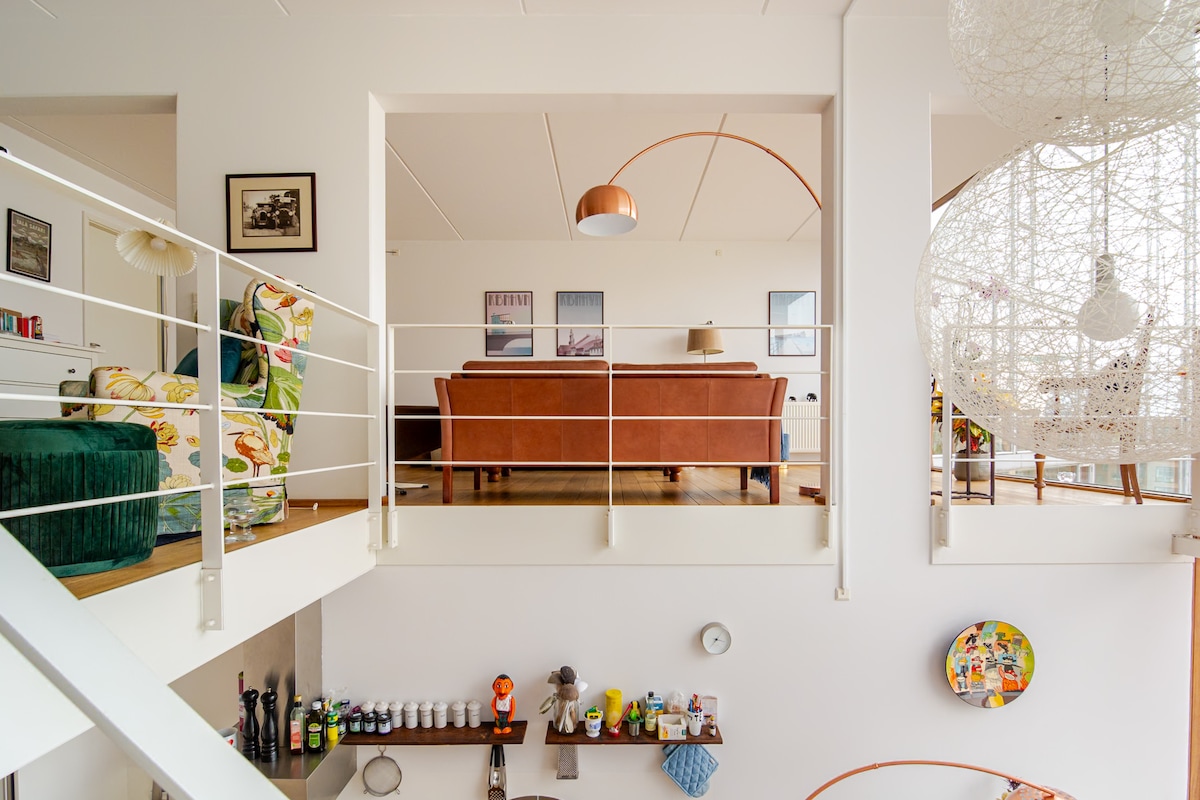 An open-plan living area showcases a mix of modern and vintage decor. A brown leather sofa is positioned across from a colorful armchair. A floor lamp provides soft lighting, while decorative items are displayed on a shelf below. Large windows allow natural light to fill the space.