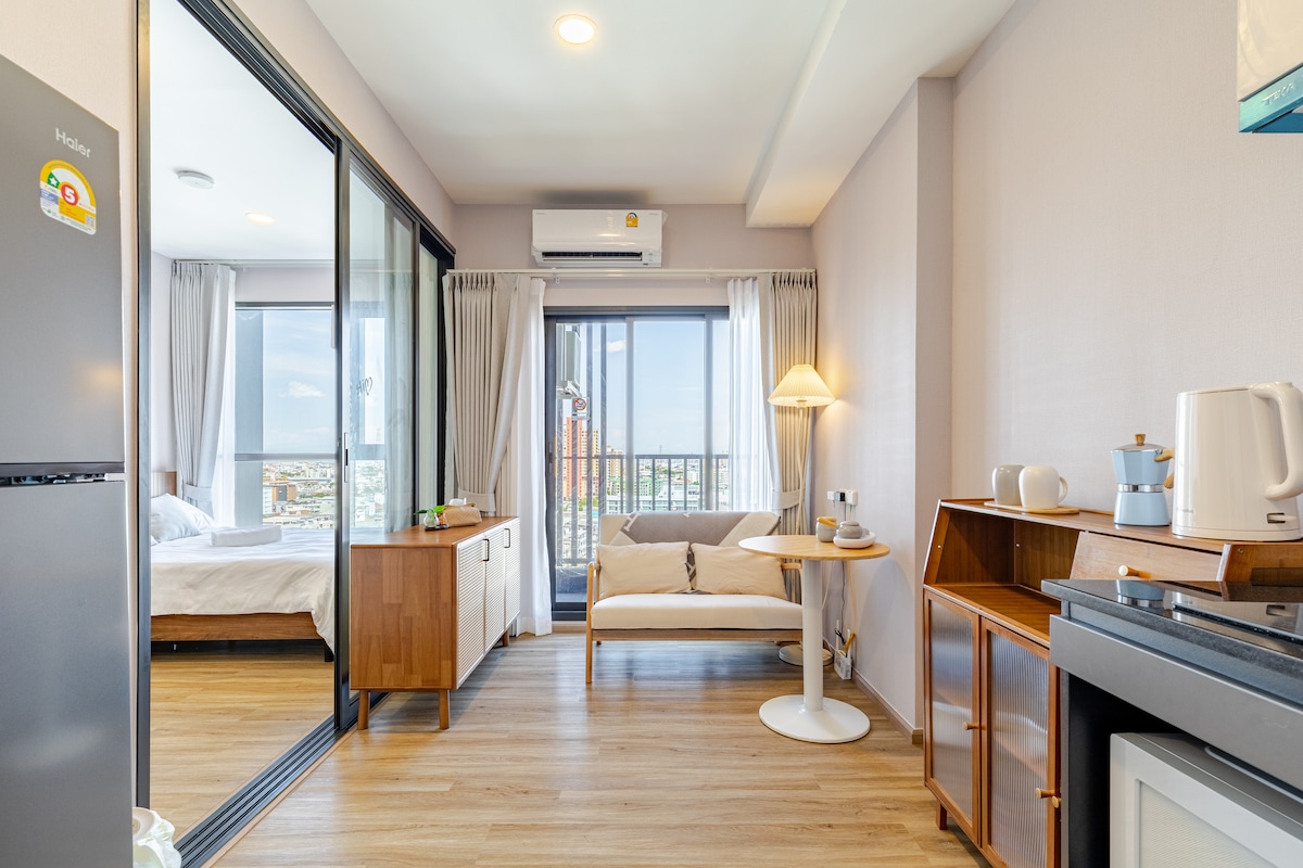A bright and airy living space features large glass sliding doors that open to a balcony. A cozy seating area is depicted with a sofa and a small table. Kitchen appliances and utensils are arranged neatly on the counter, contributing to a functional atmosphere.