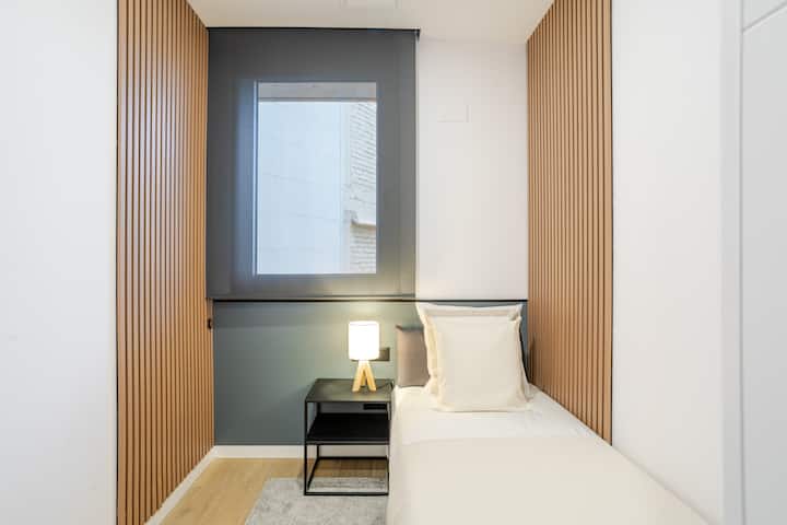 Kompaktna spavaća soba s udobnim krevetom, bijelom posteljinom i noćnim ormarićem s lampom. Drvena ukrasna ploča, ugrađeni ormar i prozor sa sjenilom stvaraju svijetao, moderan i dobro organiziran prostor.