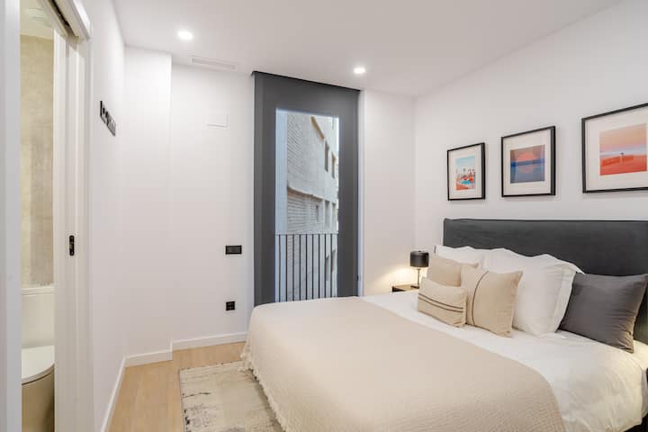 Svijetla spavaća soba s udobnim bračnim krevetom, posteljinom neutralnih boja i uokvirenim umjetničkim djelima iznad uzglavlja. Veliki prozor unosi prirodno svjetlo, naglašavajući čist dizajn, svijetle drvene podove i miran, moderan ugođaj.