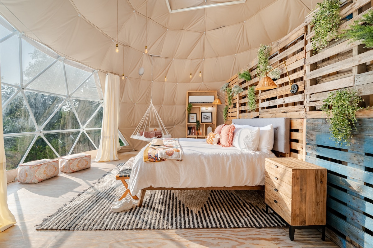 A spacious bedroom features a King-sized bed with plush bedding, framed by an artistic wall of wooden pallets. Natural light floods through large geometric windows, illuminating a cozy seating area with a hammock and decorative cushions. A bedside table adds functionality to the inviting space.
