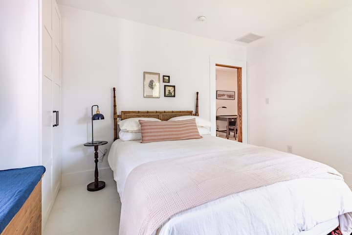 Cama tamaño queen cubierta con sábanas de algodón 100 %, un edredón de plumas alternativo y funda nórdica de algodón, con 4 almohadas.  Hay una pequeña mesa auxiliar y una lámpara. La habitación tiene una gran ventana que da al patio trasero y una ventana que da al patio lateral con un asiento de ventana.