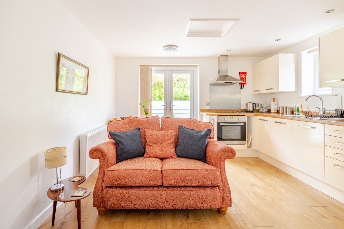 A living space features a cozy orange sofa with blue cushions, positioned in front of a well-equipped kitchen. Natural light fills the room through glass doors that lead outside. The warm wooden flooring adds to the comfortable atmosphere, complemented by wall art and a small side table.