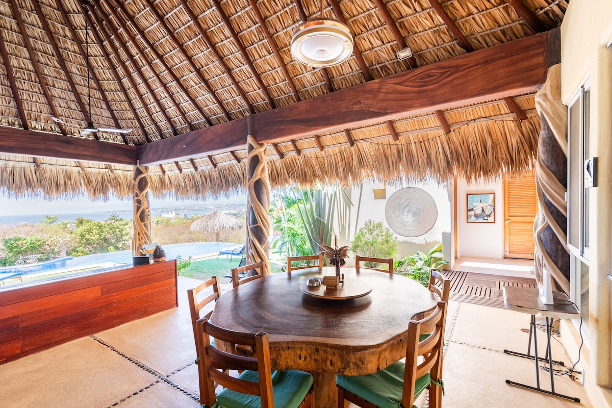 An outdoor dining area features a large circular wooden table surrounded by green upholstered chairs. The thatched roof offers shade, and glass doors open to the garden and pool view. Artwork and decorative elements enhance the natural ambiance of the space.
