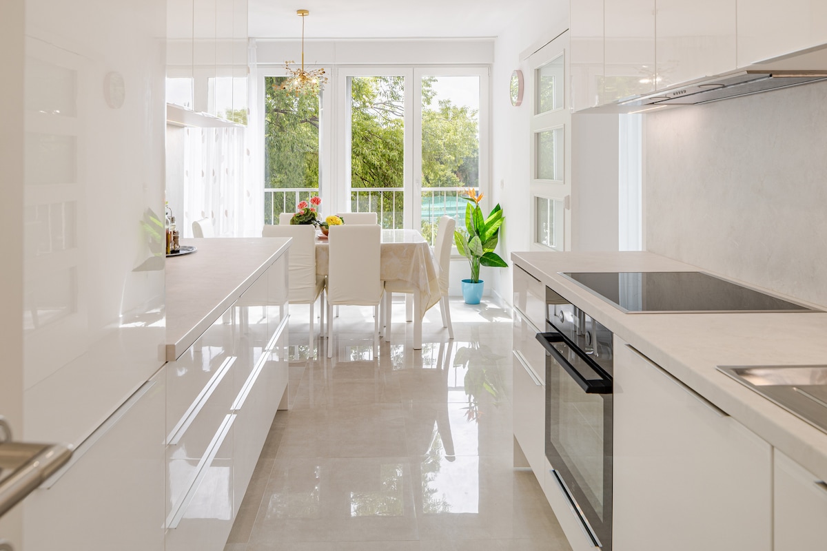 A modern kitchen is presented with sleek white cabinetry and a stone countertop. A view into the dining area reveals a table set for four, with natural light streaming in from large glass doors leading to the balcony. A potted plant adds a touch of greenery.
