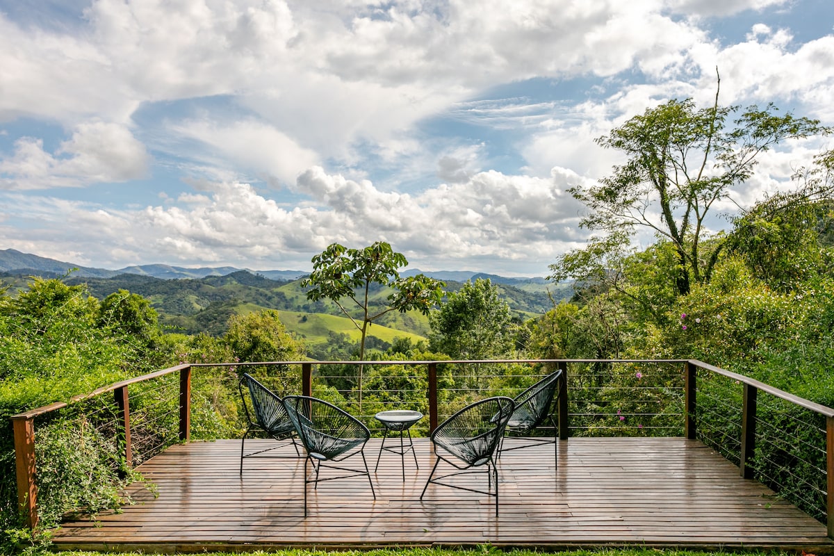 A wooden deck is equipped with two black wire chairs and a small table, offering an unobstructed view of the surrounding hills and lush greenery. The sky is scattered with clouds, enhancing the serene atmosphere.