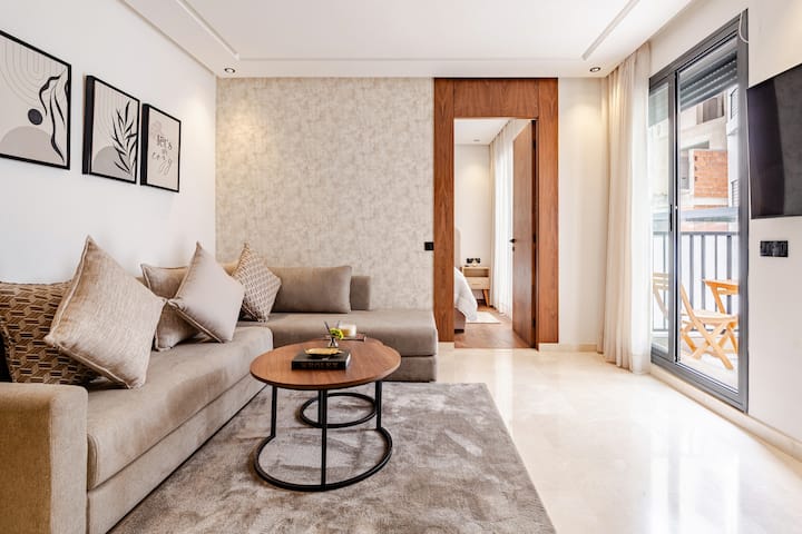 Teplá, minimalistická obývacia izba: pohodlná rohová pohovka, vankúše v pieskových odtieňoch, rozkladacie konferenčné stolíky a veľký koberec. Štruktúrovaná stena s drevenými prvkami. Okno od podlahy až po strop na balkón prináša veľkorysé prirodzené svetlo.
