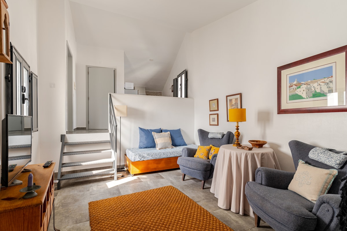 A living area features a cozy seating arrangement with a blue sofa and two armchairs. A round table is positioned in the center, adorned with a lamp. Artworks are displayed on the walls, complemented by a warm carpet underfoot. A staircase is visible in the background.