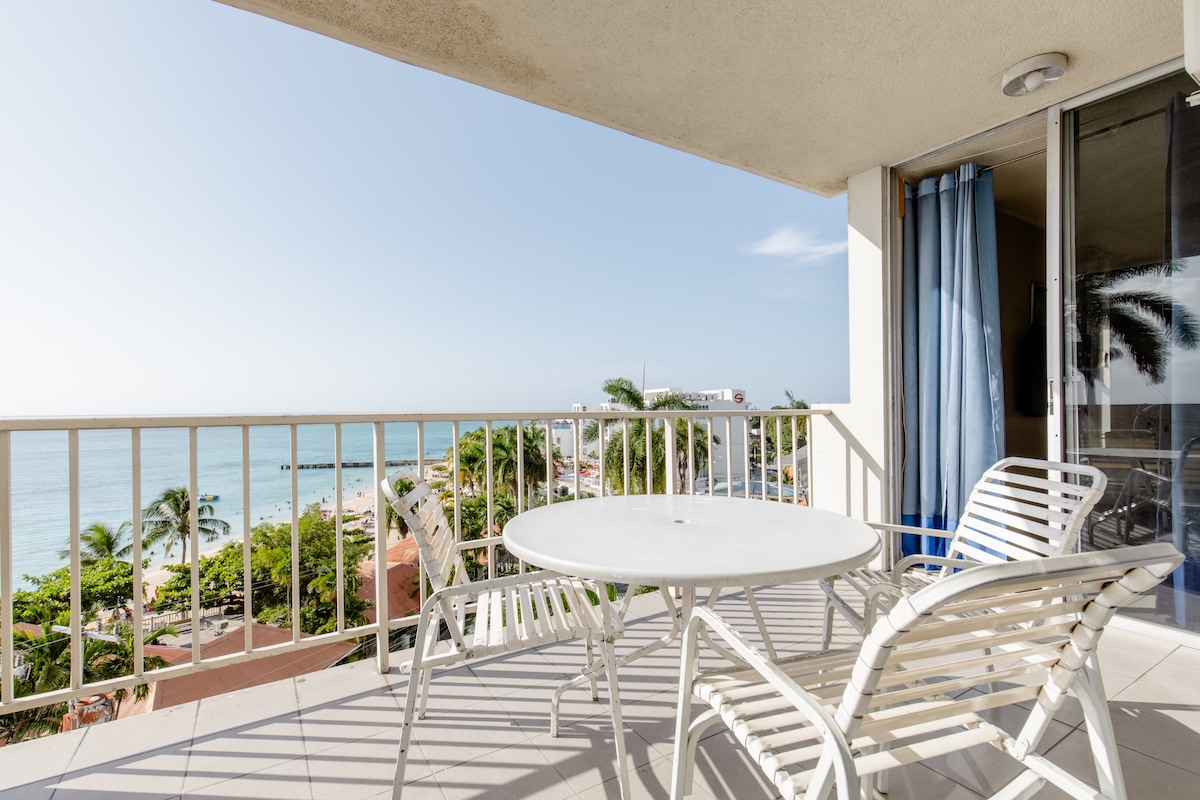 A balcony is presented with a round white table surrounded by four sleek chairs. The railing offers an uninterrupted view of the ocean and coastline, with palm trees visible in the foreground, allowing natural light to fill the space.
