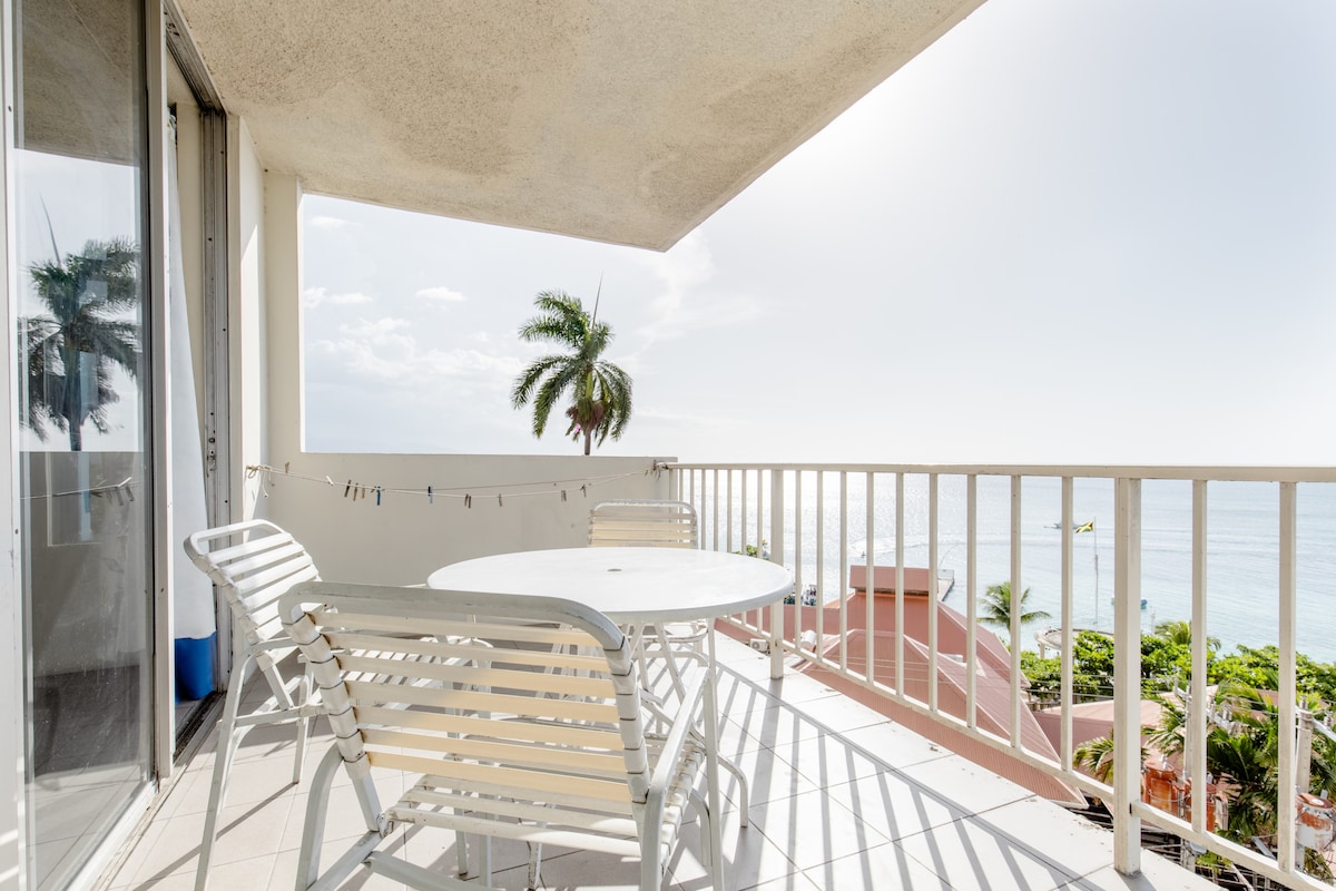 A private balcony features a round table surrounded by four chairs, with an expansive view of the ocean and palm trees. Natural light brightens the space, highlighting the tiled floor and railing, creating an inviting outdoor area for relaxation.
