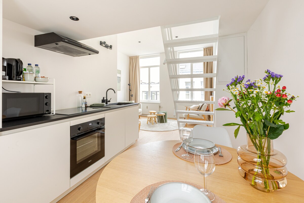 A modern kitchen area features sleek cabinetry, a built-in oven, and a microwave. A circular wooden dining table is set with plates and glasses, surrounded by white chairs. Natural light floods the space through a large window, highlighting a vase of fresh flowers on the table.