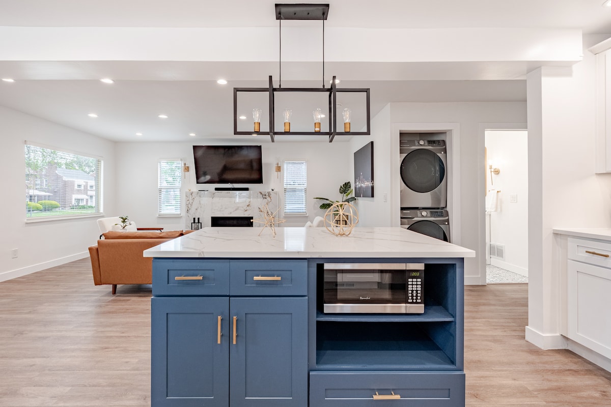 The open-concept living space features a modern kitchen island with a blue base and white countertop, including an under-counter microwave. Natural light fills the area through multiple windows, highlighting the nearby seating and entertainment setup, with a laundry area visible in the background.