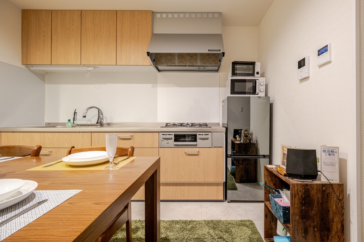 The kitchen area is equipped with wooden cabinetry and modern appliances, including a gas stove, microwave, and refrigerator. A dining table is set for a meal, with plates and glasses arranged neatly. A small workspace with a laptop is positioned near the rustic wooden shelf.