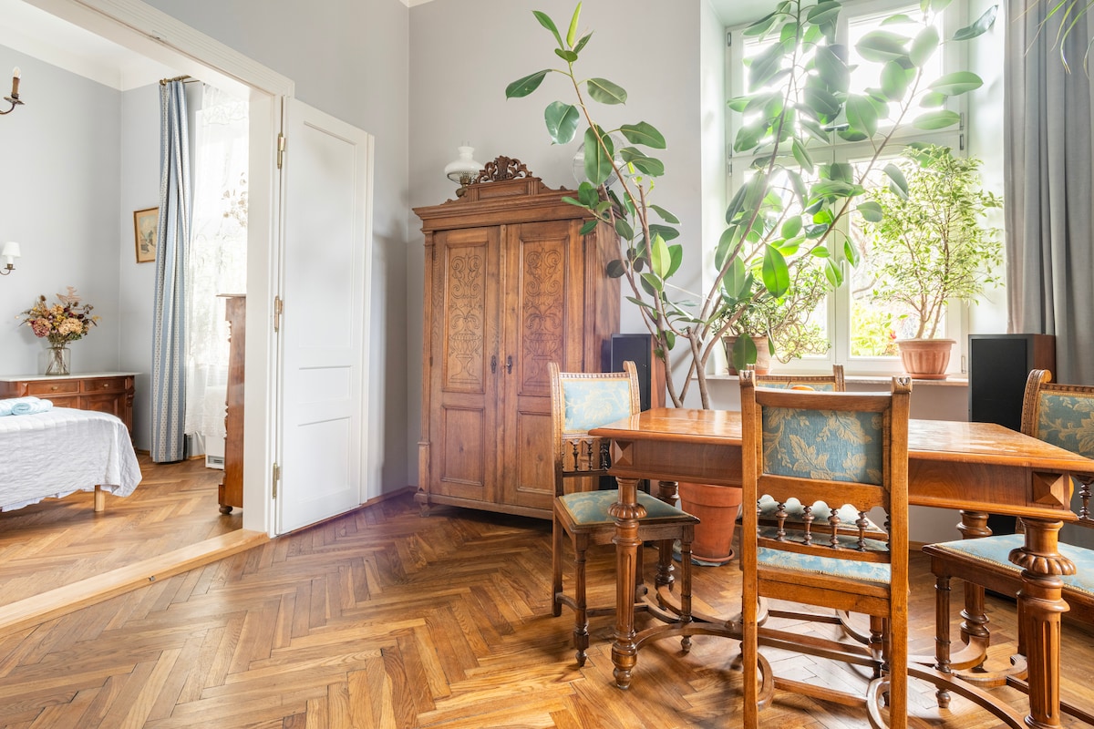 An antique dining table with wooden chairs occupies a sunlit corner, surrounded by greenery from potted plants. A vintage wooden cabinet stands nearby, while soft light filters through tall windows, illuminating the hardwood floor. The adjoining space is partially visible, revealing a cozy sleeping area.