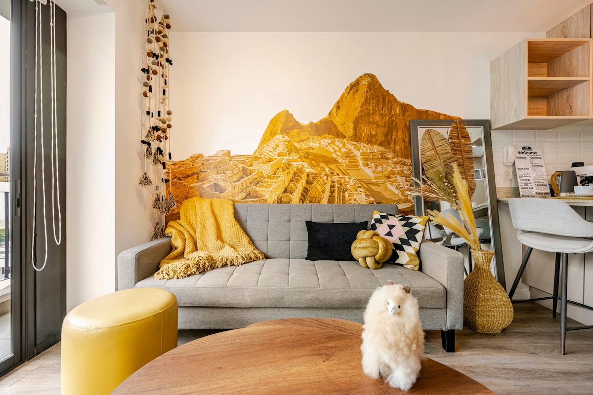 A modern living area features a grey sofa adorned with throw pillows and a warm yellow blanket. A striking mural of a mountain landscape adorns the wall, complemented by decorative elements like plants and artful textiles. Natural light enters through large windows.