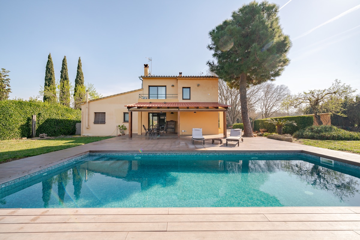 An outdoor view of a charming apartment features a private pool surrounded by a spacious deck. Lush greenery and trees provide a natural backdrop, while the structure showcases large windows and a covered patio area for relaxation. The serene setting emphasizes privacy and tranquility.