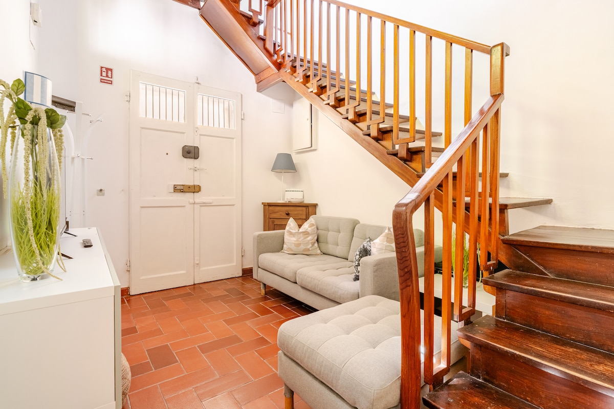A welcoming entrance area is depicted, featuring a light grey sofa and a side table. The staircase, constructed from warm wood, elegantly ascends to the upper level. Natural light enters through the front door, highlighting the terracotta tile flooring.