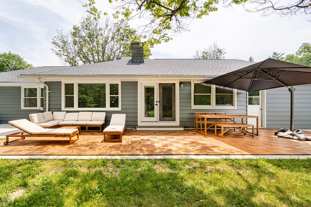 A spacious outdoor deck features a sectional lounge sofa alongside a dining area with a wooden table. Large windows provide views into the house. Sunlight filters through nearby trees, creating a relaxed outdoor setting.