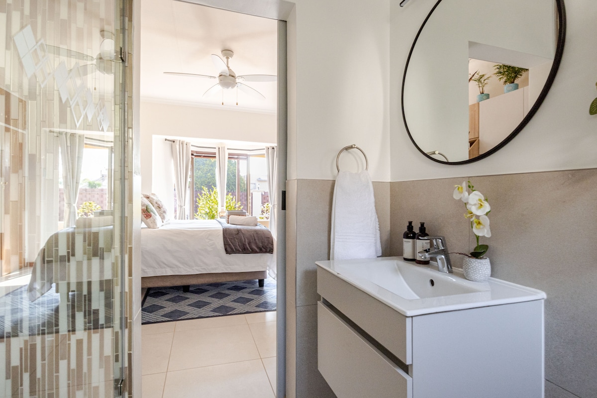 A modern en-suite bathroom features a sleek white vanity with a circular mirror above. A soothing colour palette is complemented by natural light streaming into a spacious bedroom visible through a doorway, showcasing a comfortable bed and soft furnishings.
