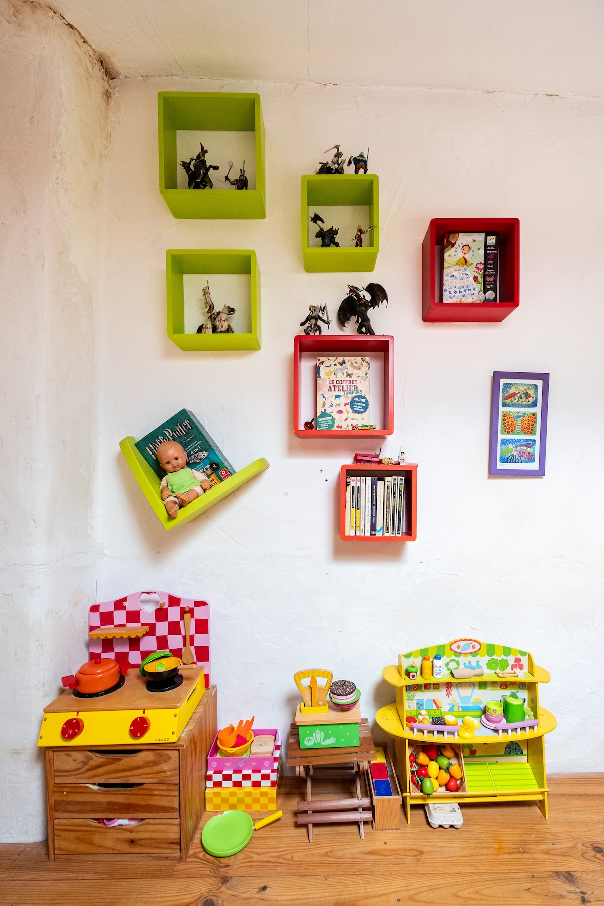 A vibrant play area features colorful shelving with assorted toys and books organized in open cubes. Below, a wooden kitchen set is accompanied by various cooking utensils and food items. The warm wooden floor complements the playful arrangement, inviting family activities.