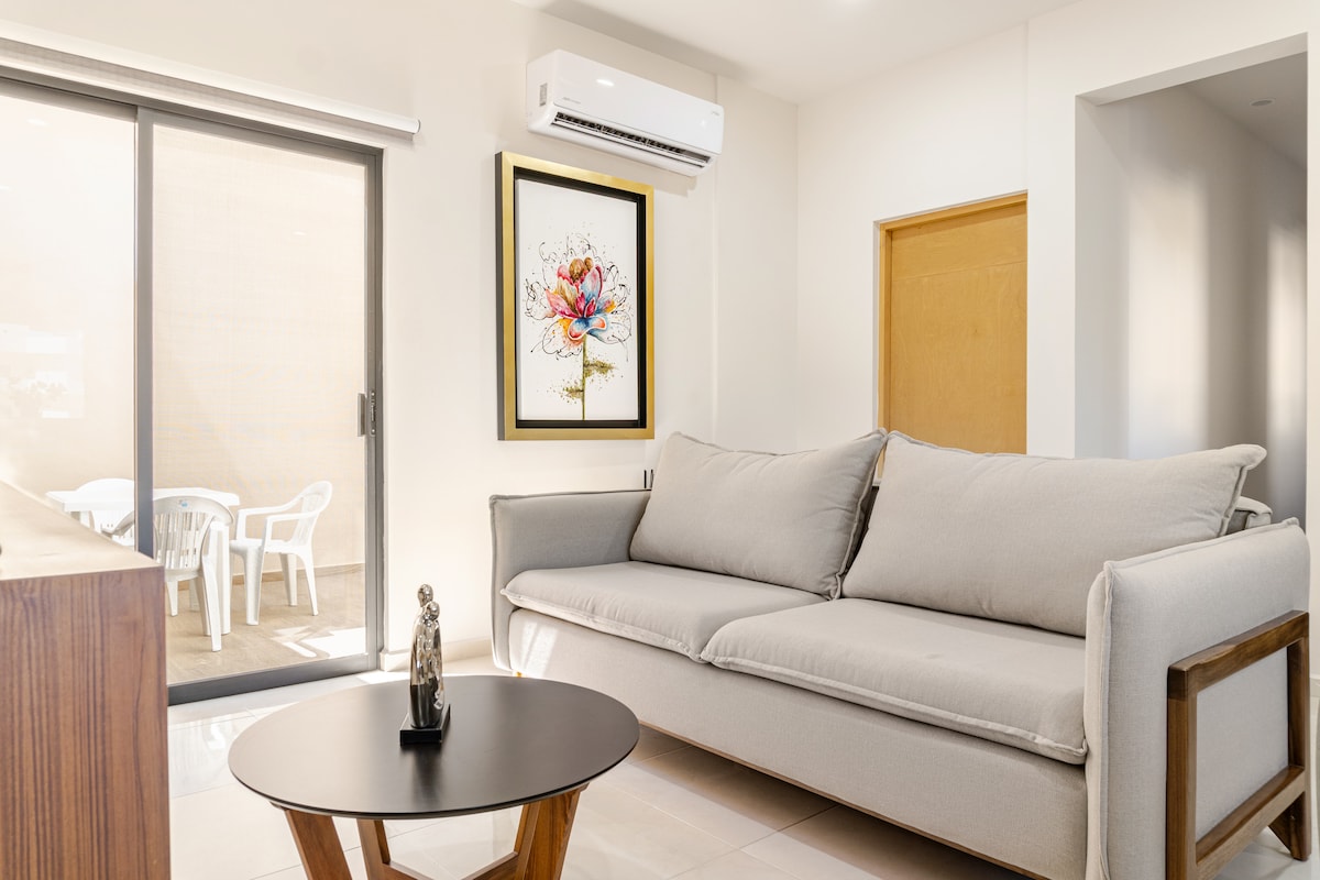 A light-filled living area is furnished with a spacious gray sofa and a black coffee table. A piece of wall art featuring a floral design adds a pop of color. Glass sliding doors lead to an outdoor patio, enhancing the open space.